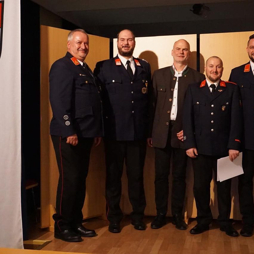 Five men in military uniforms stand together in a room. One holds a piece of paper, another wears a medal. They are likely celebrating a special occasion.