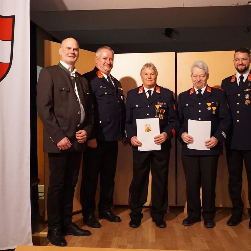 Five men in uniform are standing in a row. The two in the middle are holding certificates. Behind them is a flag with a logo.