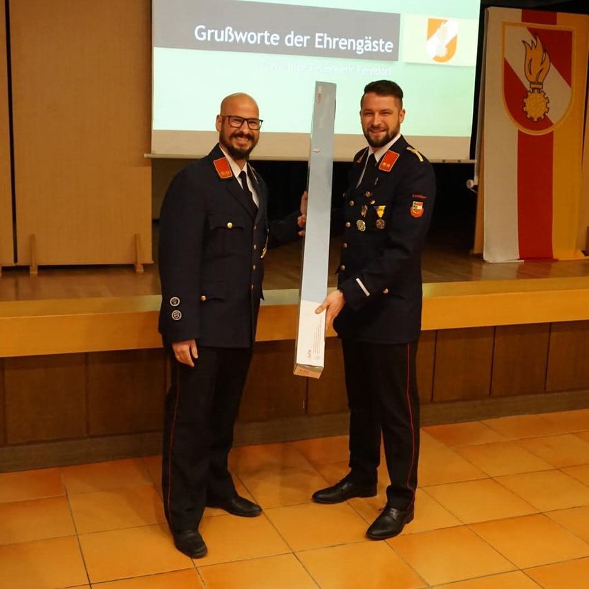 Two men in military uniforms hold a white box while standing in front of a stage with a projector screen showing 'Grußworte der Ehrengäste'.