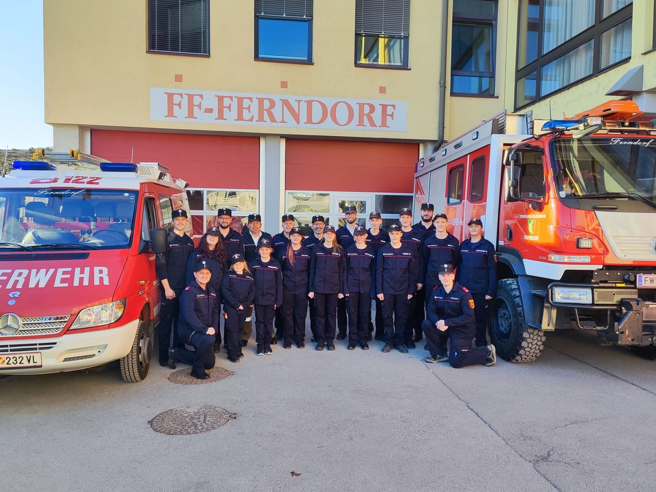 Eine Gruppe Feuerwehrleute in Uniform posiert für ein Foto vor ihrer Feuerwache, wobei im Hintergrund zwei Feuerwehrfahrzeuge zu sehen sind.