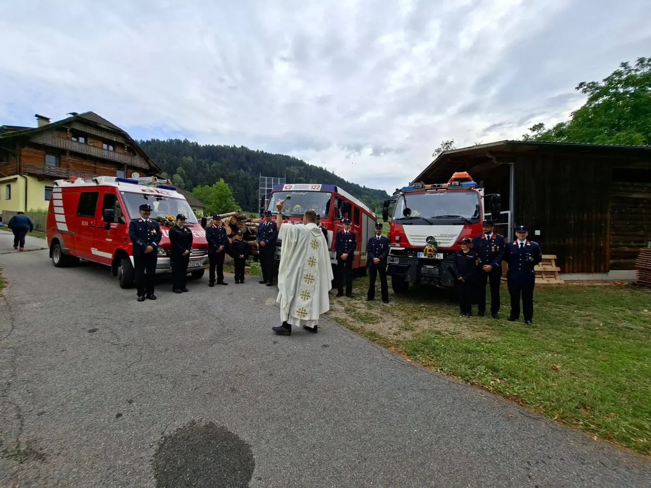 Ein Priester in einer weißen Robe steht vor einer Gruppe von Feuerwehrleuten in Uniform, mit geparkten Feuerwehrwagen hinter ihnen an einem sonnigen Tag.