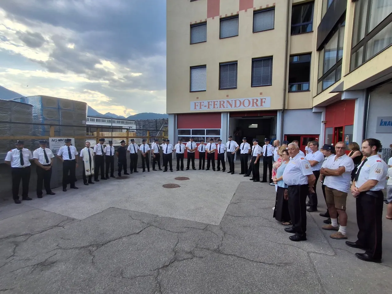Eine Gruppe von Feuerwehrleuten und Zivilisten steht in einem Kreis vor der FF-Ferndorf Feuerwehrstation. Die Feuerwehrleute sind in Uniform und die Zivilisten beobachten.