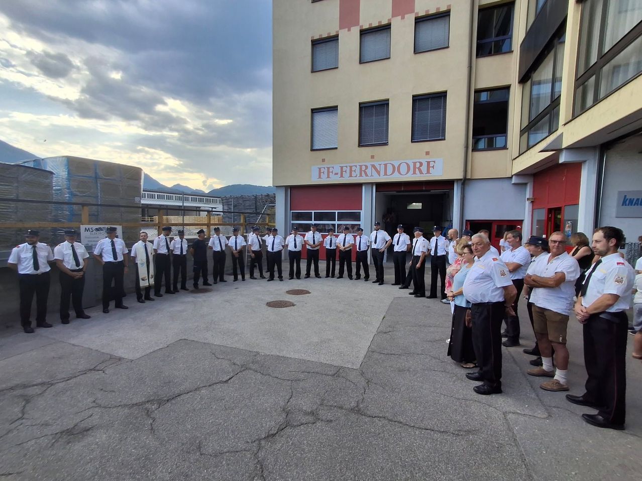 Eine Gruppe von Feuerwehrleuten und Zivilisten steht in einem Kreis vor der FF-Ferndorf Feuerwehrstation. Die Feuerwehrleute sind in Uniform und die Zivilisten beobachten.