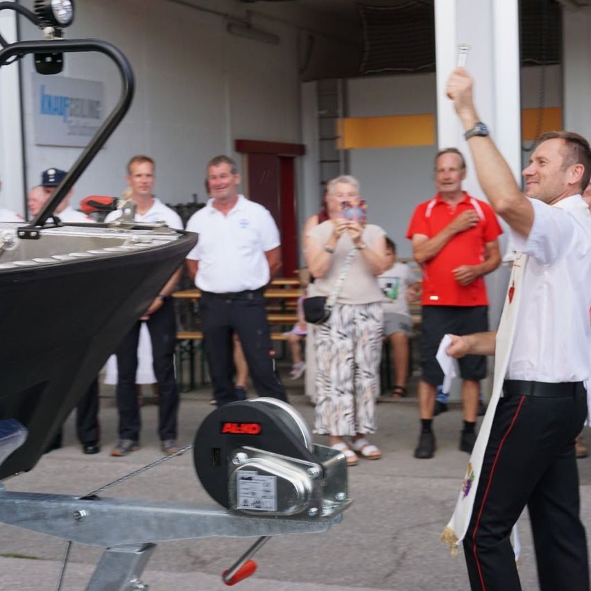Eine Gruppe von Menschen steht um ein Boot herum, ein Mann hebt die Hand, während andere Kameras halten und klatschen. Eine Frau in der Mitte macht ein Foto.