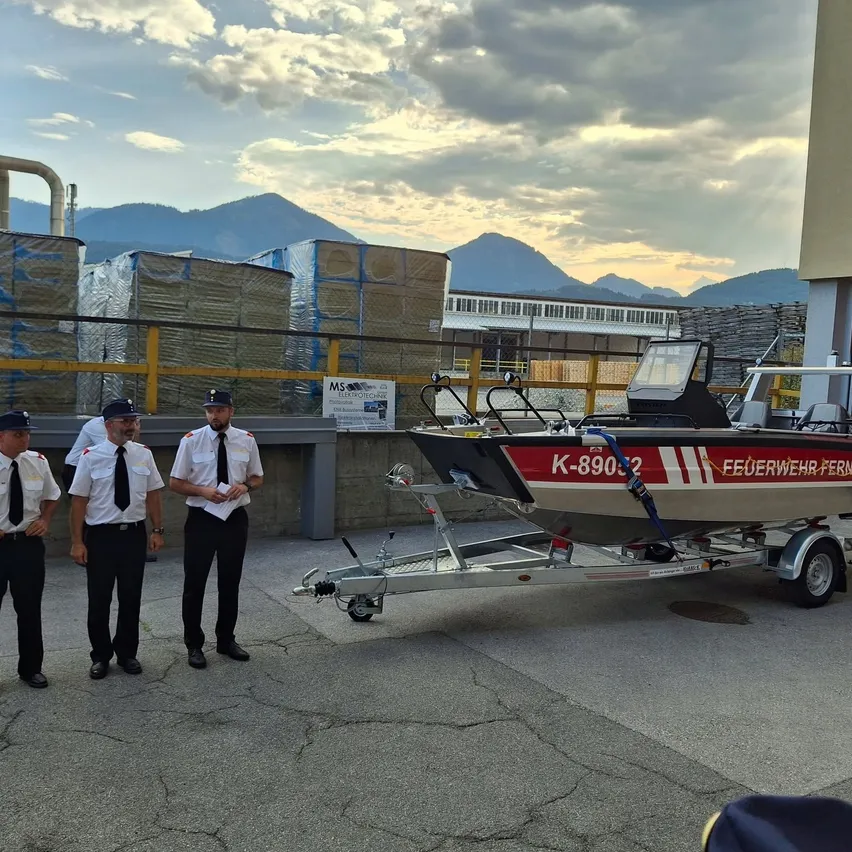 Drei Feuerwehrleute in Uniform stehen neben einem roten und weißen Boot auf einem Anhänger, mit Bergen und einem bewölkten Himmel im Hintergrund.