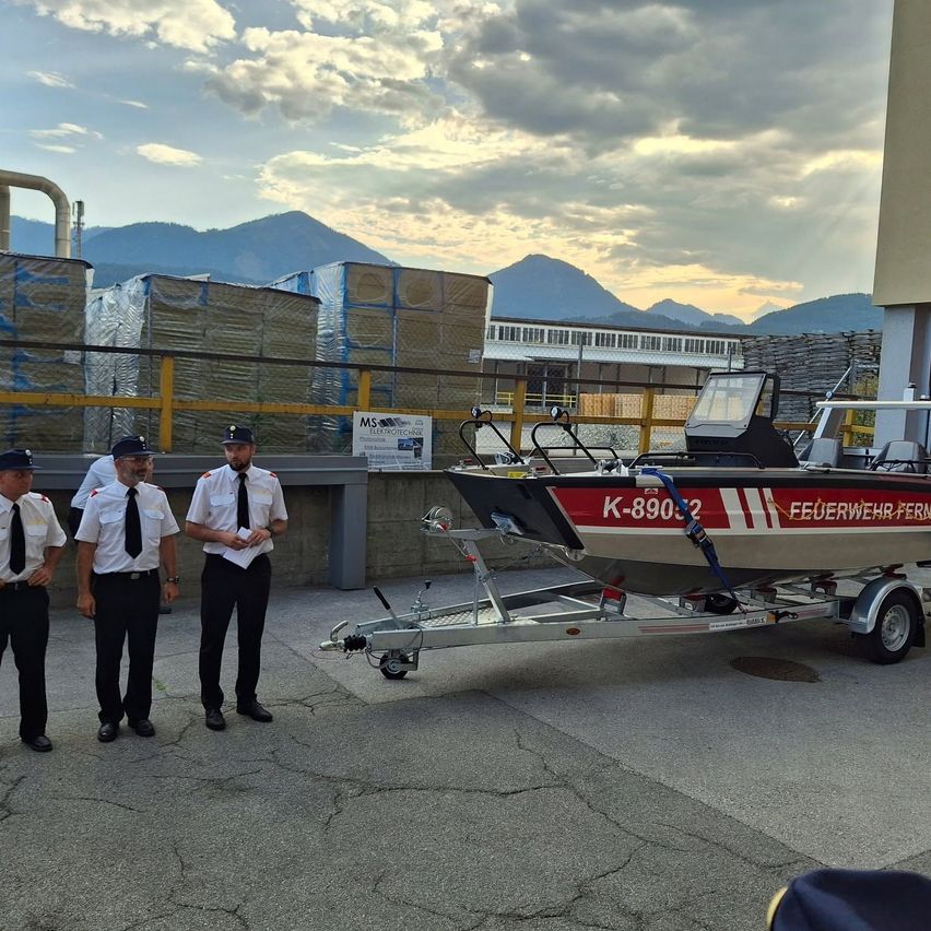 Drei Feuerwehrleute in Uniform stehen neben einem roten und weißen Boot auf einem Anhänger, mit Bergen und einem bewölkten Himmel im Hintergrund.