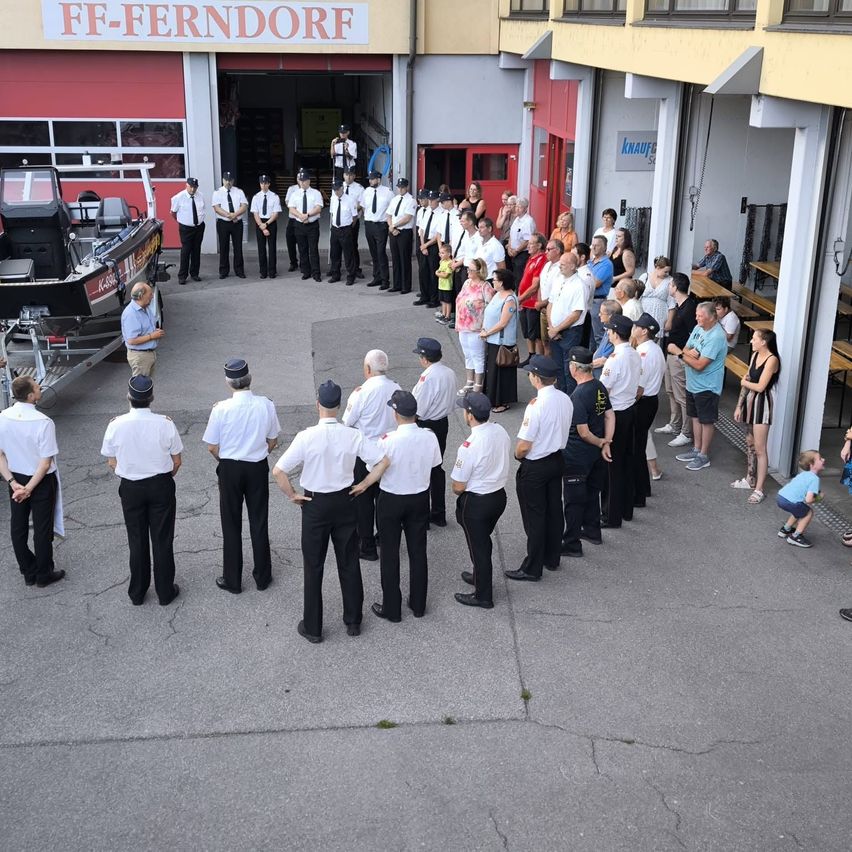 Eine Gruppe uniformierter Personen steht in einem Kreis vor einem Gebäude mit dem Schild FF-Ferndorf. Dahinter ist ein Boot geparkt, und mehrere Personen stehen in der Nähe von Bänken.