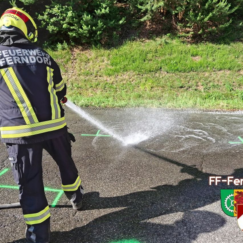 Ein Feuerwehrmann in gelber und schwarzer Uniform sprüht Wasser aus einem Schlauch auf einen asphaltierten Weg. Grüne Markierungen sind auf dem Boden.