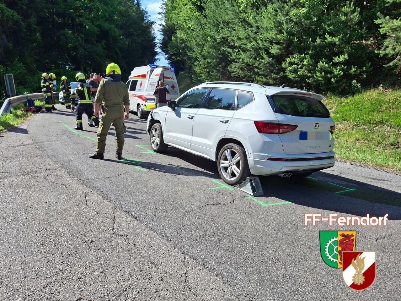 Einsatzkräfte umgeben einen weißen SUV mit einem platten Reifen am Straßenrand, mit einem Krankenwagen im Hintergrund.