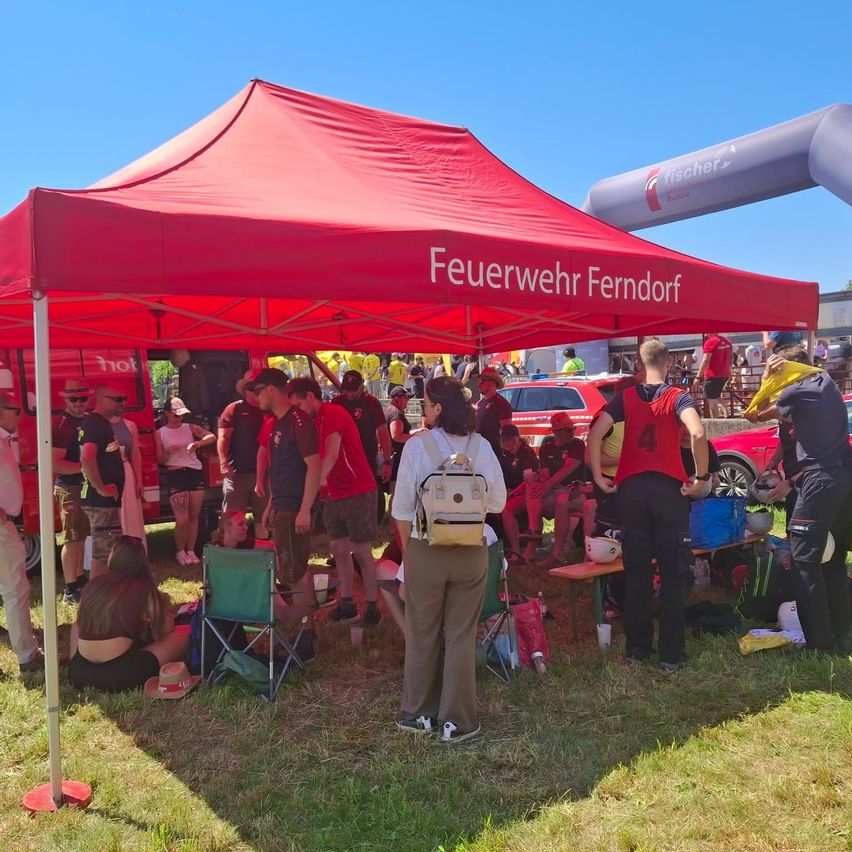 Menschen versammeln sich unter einem roten Zelt mit der Aufschrift 'Feuerwehr Ferndorf'. Einige sitzen auf Stühlen, andere stehen. Ein Feuerwehrauto ist in der Nähe.