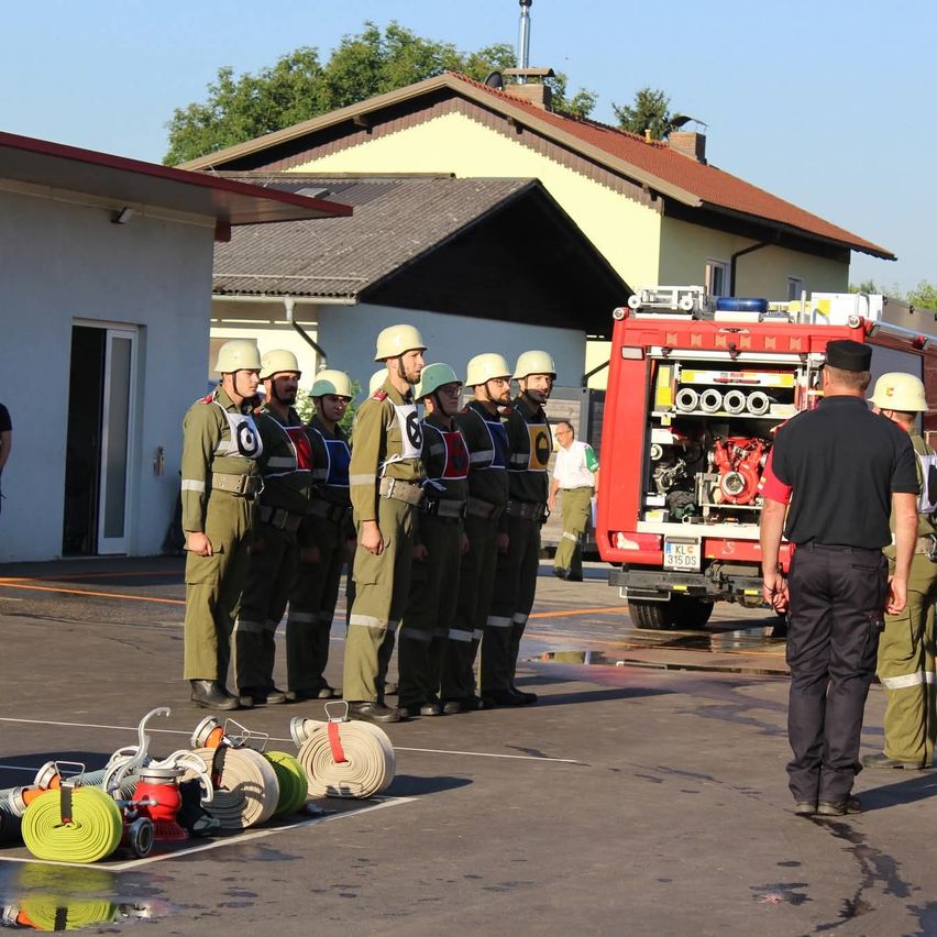 Sechs Feuerwehrleute in Uniform stehen in einer Reihe vor einem Gebäude. Ein Feuerwehrwagen ist in der Nähe geparkt, und ein Mann in einem schwarzen Hemd beobachtet sie.