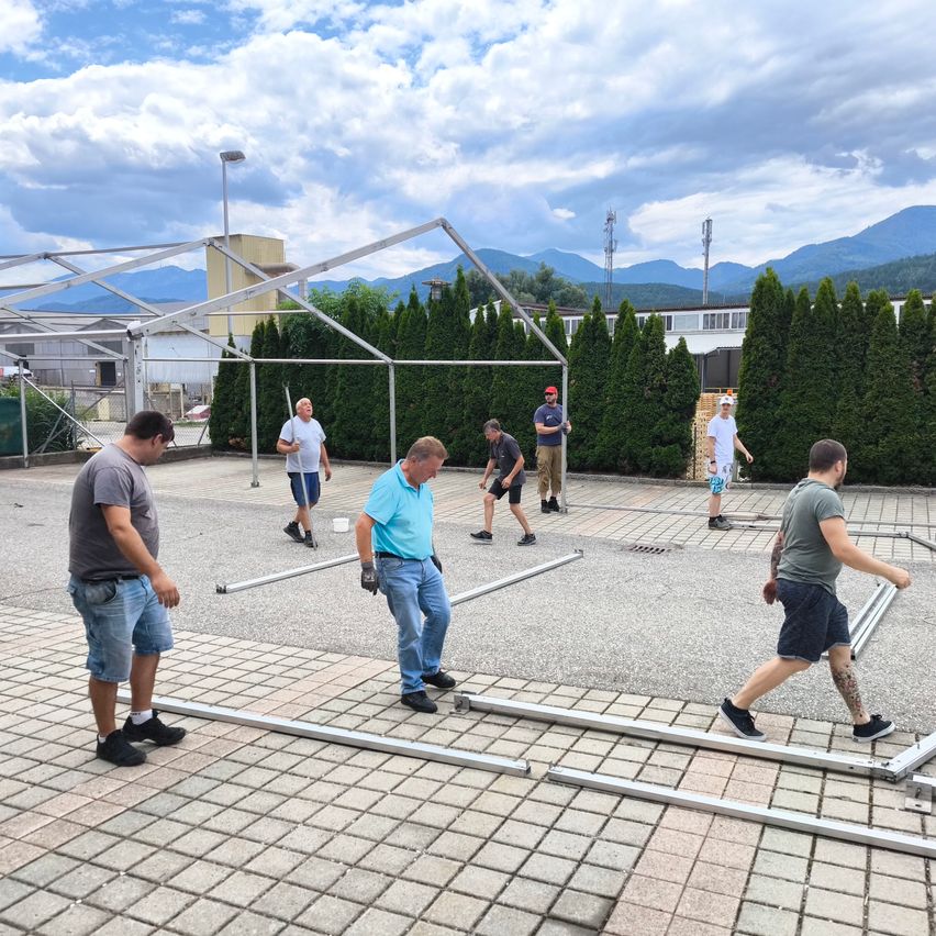 A group of people are working on a metal structure in an outdoor area with mountains in the distance. They are wearing casual clothes and some are wearing gloves. They are standing on a brick floor and seem to be fixing something. In the distance, there is a tall metal fence with a building behind it.