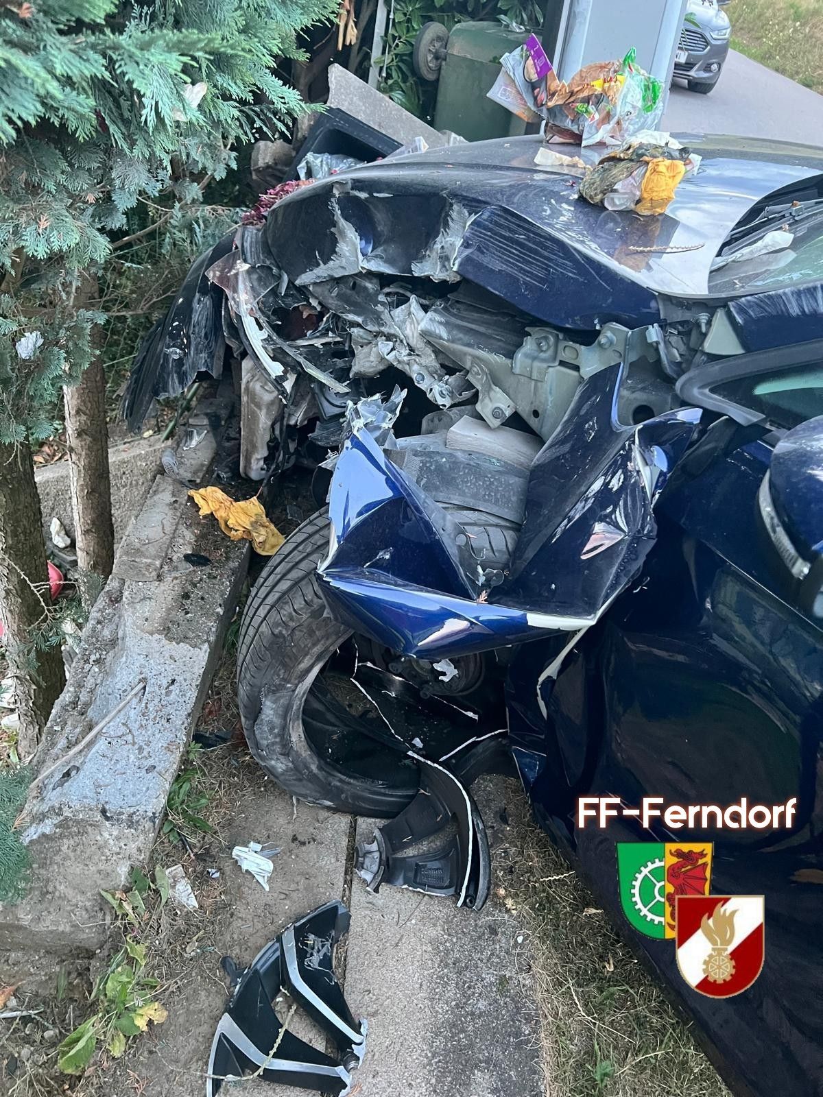 A severely damaged blue car is wrecked on the side of the road, with debris scattered around and a tree nearby.