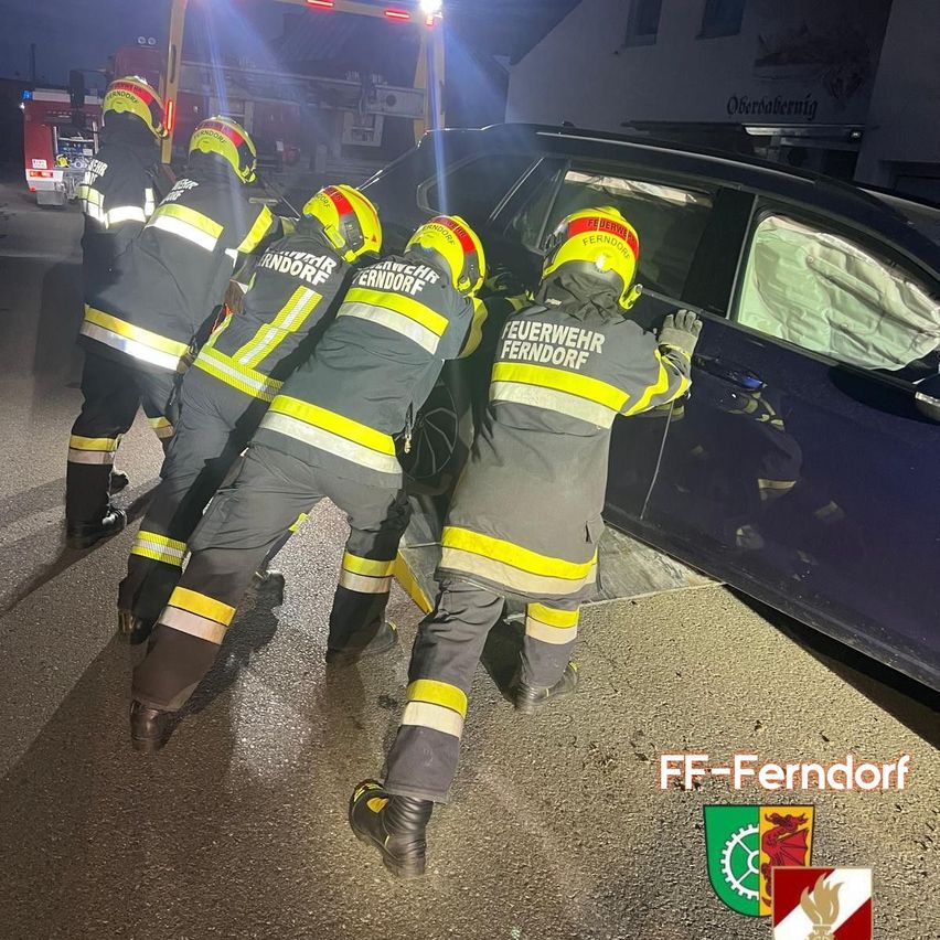 Five firefighters in protective gear are working together to open a damaged car door at night.