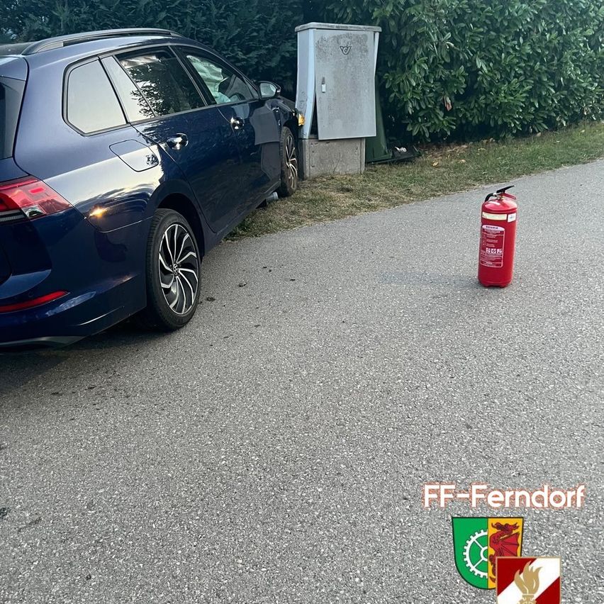 A blue car is parked on the side of the road with a fire extinguisher nearby. The car is parked on a gravel surface with a metal box behind it.
