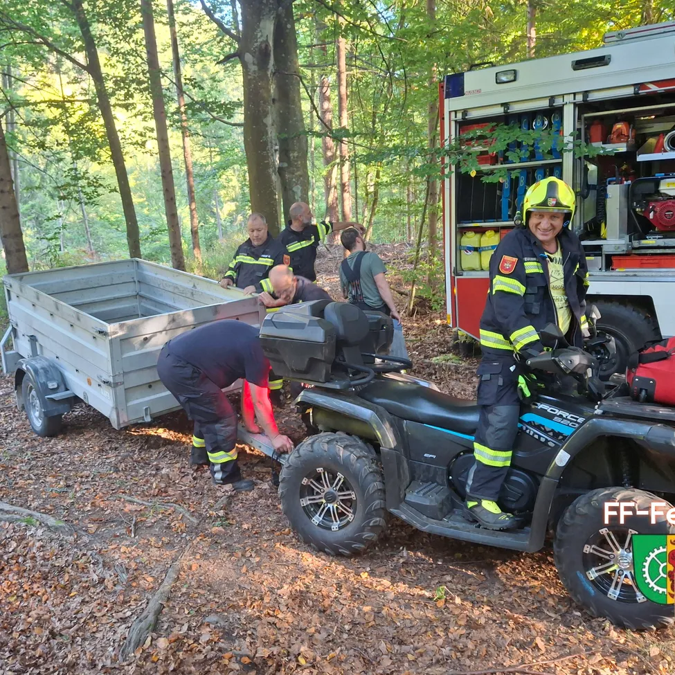 Eine Gruppe von Feuerwehrleuten arbeitet an einem schwarzen ATV in einem Wald. Eine Person sitzt auf dem ATV und eine andere repariert etwas am Anhänger. Ein Feuerwehrwagen ist hinter ihnen geparkt.