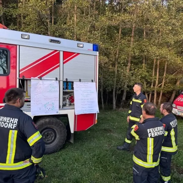 Eine Gruppe von Feuerwehrleuten ist um einen Feuerwehrwagen versammelt. Sie diskutieren etwas, während sie ein Blatt Papier betrachten.