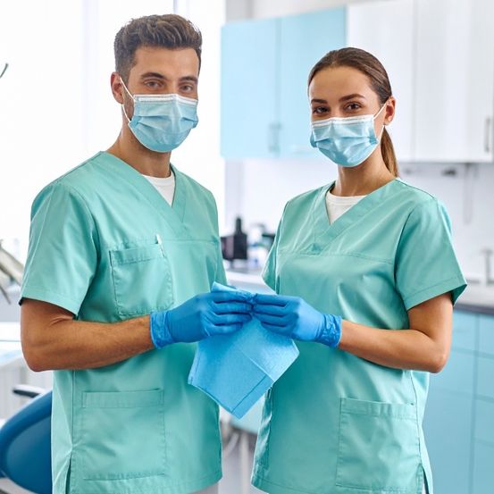 Zwei Gesundheitspersonal in türkisen Kitteln und Gesichtsmasken stehen in einer Klinik. Einer hält ein blaues Taschentuch, beide schauen etwas an. Dahinter steht ein Zahnarztstuhl und Schränke.