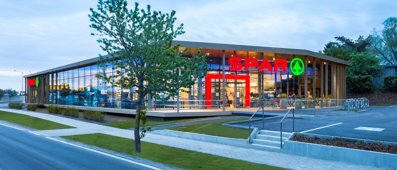 Das Äußere eines modernen Spar-Supermarkts mit Glaswänden, einem roten Eingang und einem grünen Logo. Ein Baum steht vor dem Gebäude.