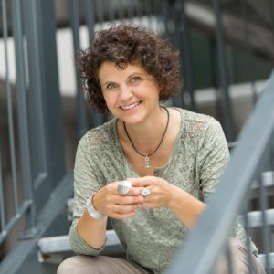 Eine Frau mit lockigem Haar sitzt auf einer Treppe, lächelt und hält eine Tasse in den Händen.