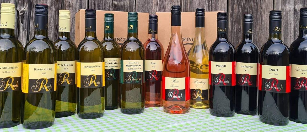 Eine Sammlung von Ribisch-Weinen, die vor einer Holzwand ausgestellt sind. Sie sind in Reihen angeordnet, jede Flasche ist mit ihrer Art und dem Jahrgang beschriftet.