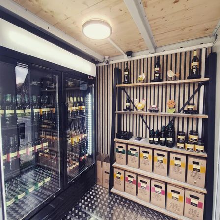 Ein kleiner Kühlraum mit Glastür und Regalen, gefüllt mit Flaschen und Gläsern, beleuchtet von Deckenlicht, an einer gestreiften Wand.