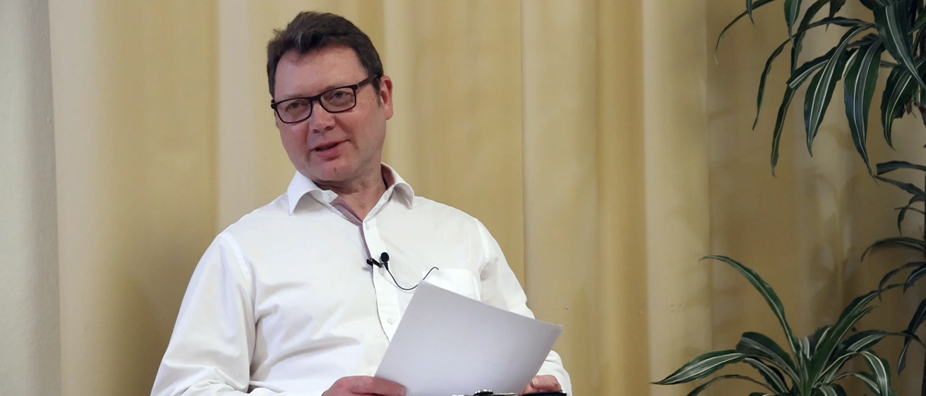 Ein Mann mit Brille sitzt in einem Stuhl und hält ein Papier in der Hand, möglicherweise hält er eine Rede.