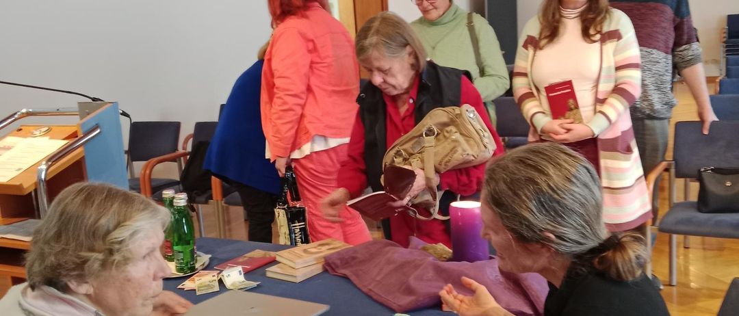 Mehrere Personen stehen um einen Tisch mit einer blauen Tischdecke herum. Eine Person hält ein Buch und scheint mit einer anderen Person zu sprechen.