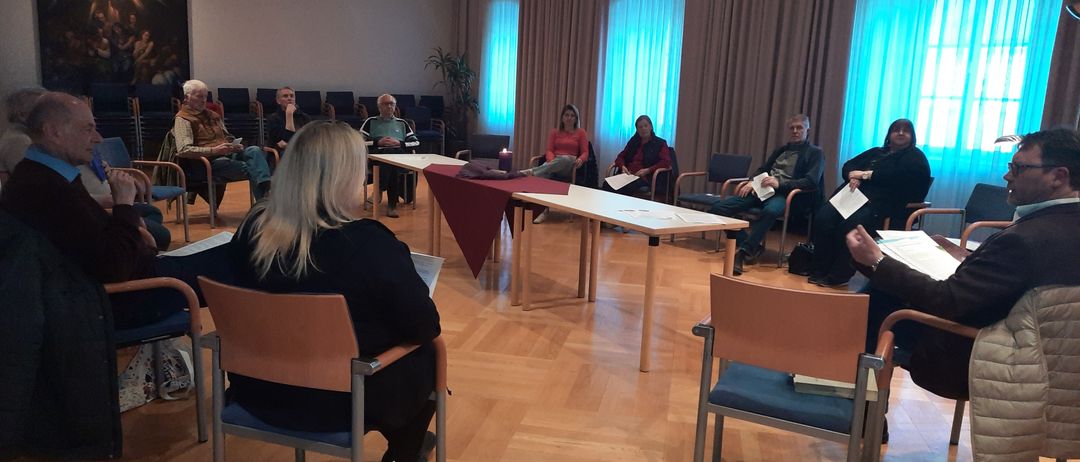 Eine Gruppe von Menschen sitzt im Kreis in einem Raum. Der Raum hat einen Holzboden und einen großen Vorhang an der Wand.