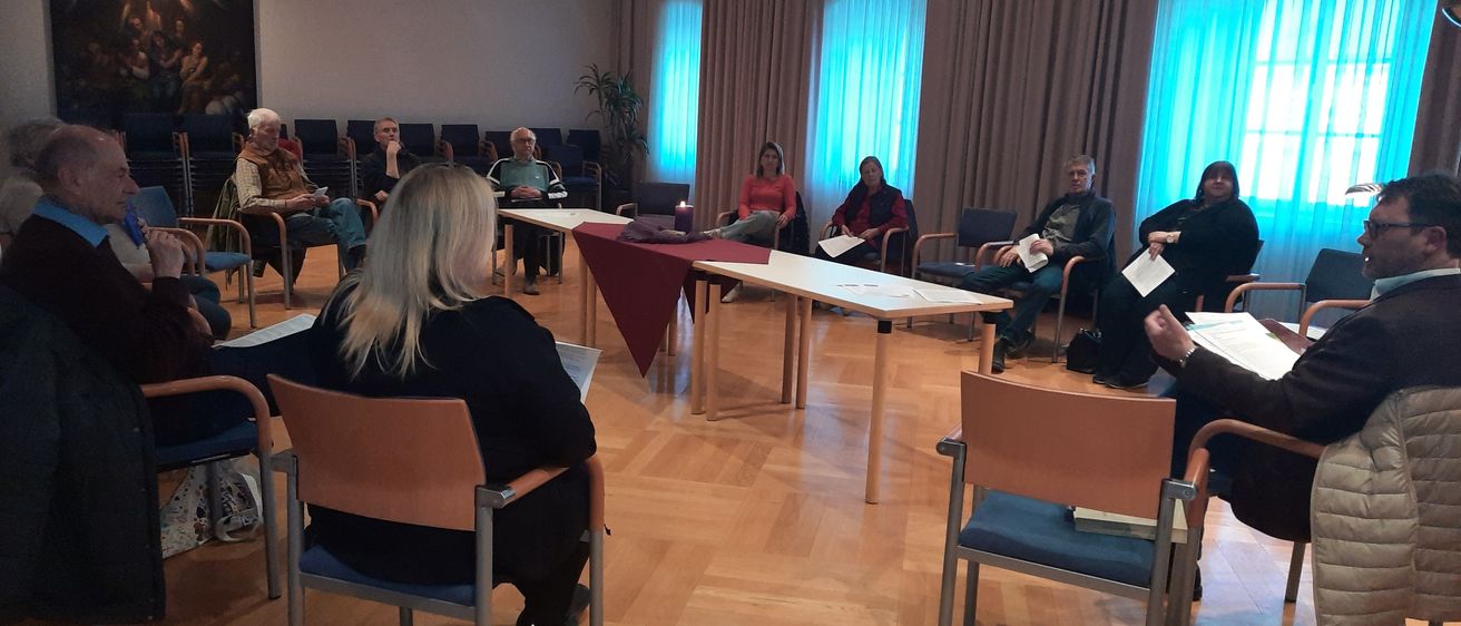 Eine Gruppe von Menschen sitzt im Kreis in einem Raum. Der Raum hat einen Holzboden und einen großen Vorhang an der Wand.