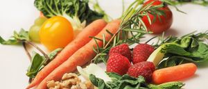 Eine Auswahl an frischem Obst und Gemüse, darunter Karotten, Tomaten, Himbeeren und Walnüsse, auf einer weißen Oberfläche präsentiert.