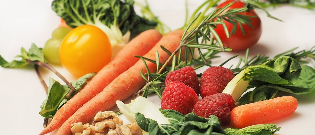 Eine Auswahl an frischem Obst und Gemüse, darunter Karotten, Tomaten, Himbeeren und Walnüsse, auf einer weißen Oberfläche präsentiert.