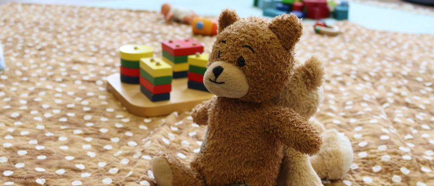Ein brauner Teddybär sitzt auf einer Tüpfeldecke mit Spielzeug wie bunten Bauklötzen darum. Ein weiterer Teddybär ist teilweise dahinter versteckt.