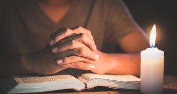 Eine Person betet mit verschränkten Händen über einem offenen Buch auf einem Tisch, eine brennende Kerze in der Nähe wirft ein warmes Licht.