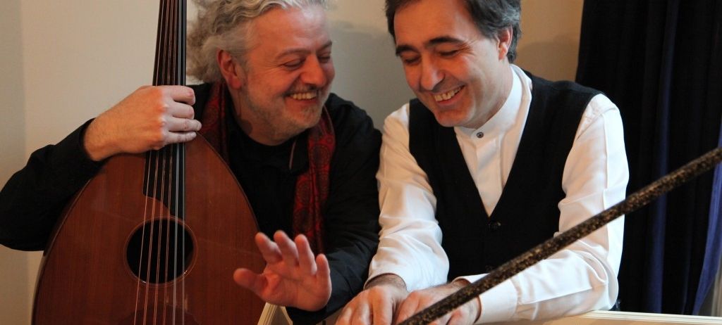 Zwei Männer lächeln und spielen Musikinstrumente. Einer spielt eine Laute, der andere ein Qanun. Sie sitzen und schauen sich an.