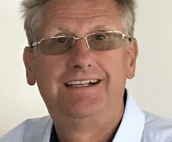 Ein Mann mit kurzen, silbrig-grauen Haaren, der eine Brille trägt, lächelt in die Kamera. Er ist in ein hellblaues Hemd mit schwarzem Kragen gekleidet.