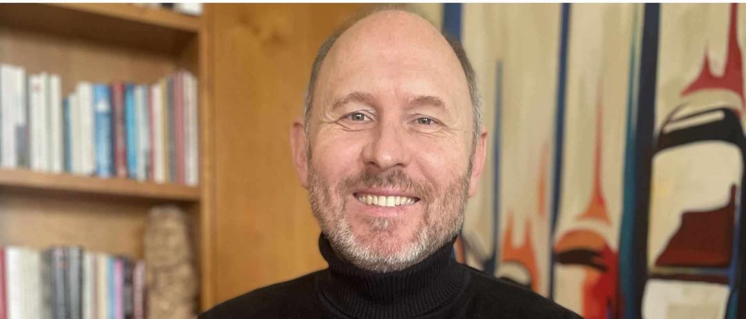 Ein Mann in einem schwarzen Rollkragen steht mit verschränkten Armen vor einem Bücherregal und einem farbenfrohen Gemälde.