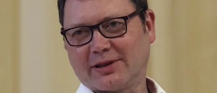 Ein Mann mit Brille und weißem Hemd lächelt und schaut nach vorne in einer Innenumgebung.