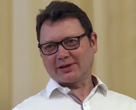 Ein Mann mit Brille und weißem Hemd lächelt und schaut nach vorne in einer Innenumgebung.