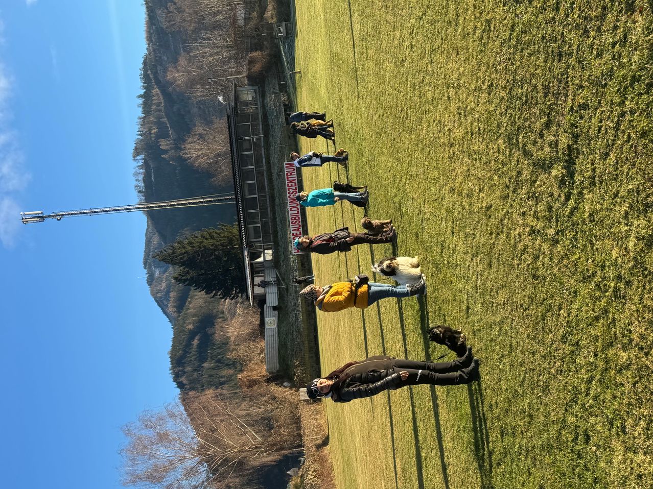 Eine Gruppe von Menschen mit Hunden steht in einer Reihe auf einem Rasenfeld, mit einem Gebäude und einem Berg im Hintergrund.
