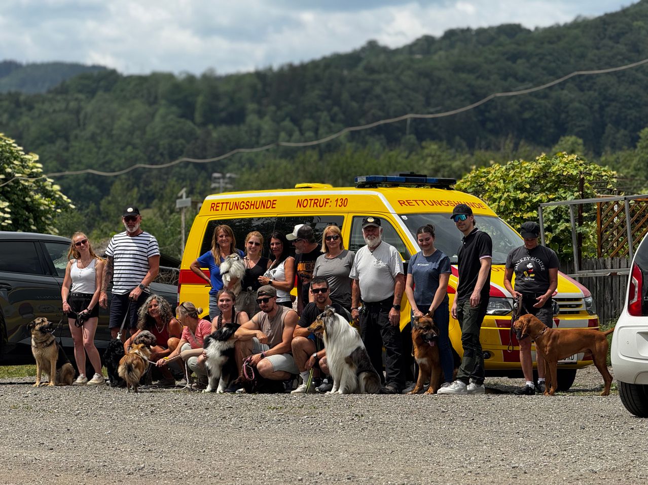 Eine Gruppe von Menschen mit Hunden steht vor einem gelben Rettungsfahrzeug. Sie tragen alle Sonnenbrillen und Hüte. Dahinter befindet sich ein Berg und eine Straße.