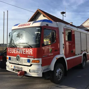 Ein roter und weißer Feuerwehrwagen steht vor einem Haus. Der Wagen hat die Aufschriften 'Adernklaa' und 'Feuerwehr'. Eine Person sitzt im Inneren des Wagens.