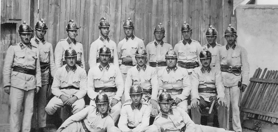 Ein Schwarz-Weiß-Foto einer Militäreinheit in Uniform, mit Helmen und Gürteln, posiert vor einem Holzzaun.