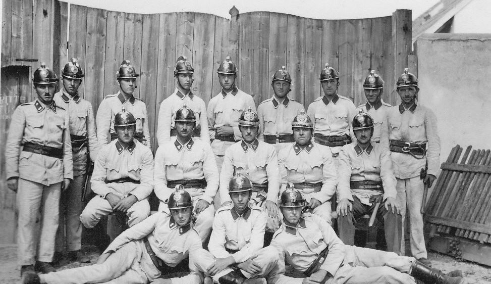 Ein Schwarz-Weiß-Foto einer Militäreinheit in Uniform, mit Helmen und Gürteln, posiert vor einem Holzzaun.