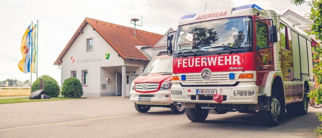 Two fire trucks are parked in front of a fire station building with a red roof. The trucks are red and white with the word 'Feuerwehr' written on the front.