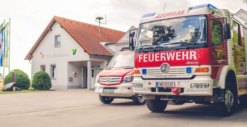 Zwei Feuerwehrfahrzeuge sind vor einem Feuerwehrhaus mit einem roten Dach geparkt. Die Fahrzeuge sind rot und weiß mit dem Wort 'Feuerwehr' auf der Front geschrieben.