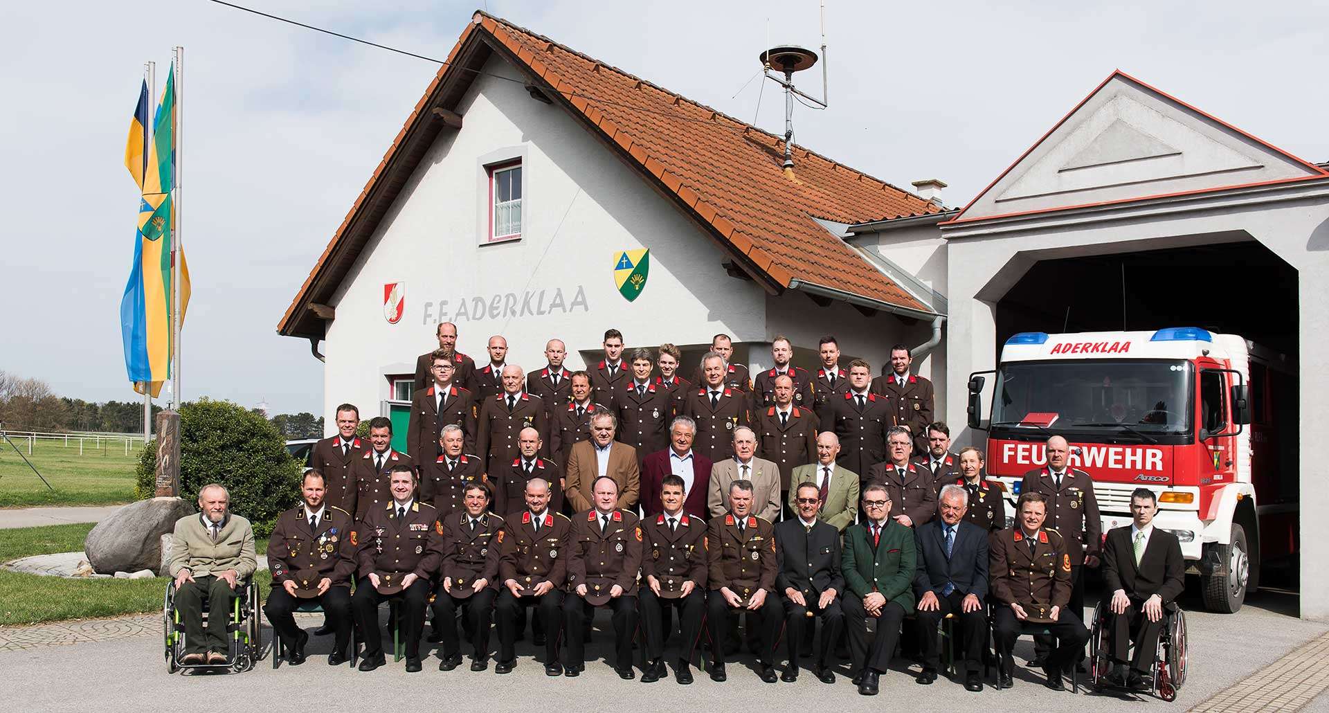Eine Gruppe uniformierter Personen, darunter einige in Militärkleidung, posiert vor einem Gebäude mit der Aufschrift 'F.Aderklaa'. Das Gebäude hat ein rotes Dach, und ein Feuerwehrauto ist rechts zu sehen.