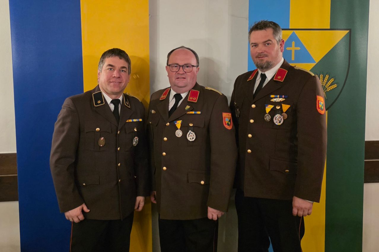 Drei Männer in Militäruniformen posieren für ein Foto. Sie tragen Medaillen und stehen vor einem blau-gelben Hintergrund.