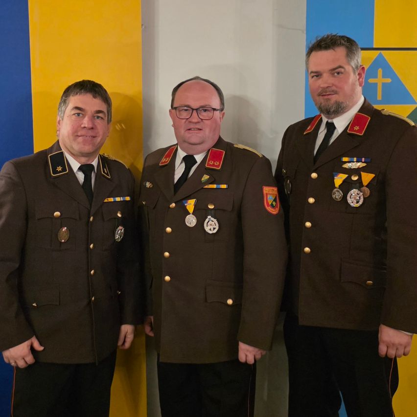 Drei Männer in Militäruniformen posieren für ein Foto. Sie tragen Medaillen und stehen vor einem blau-gelben Hintergrund.