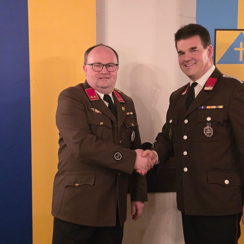 Zwei Männer in Militäruniformen schütteln sich die Hände vor einer blau-gelben Wand mit einem Symbol.